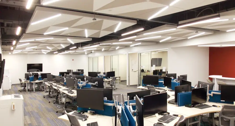 Blue Canyon Technologies' modern office space with rows of desks equipped with computer monitors, ergonomic chairs, and blue dividers. The ceiling features geometric panels with bright LED strip lighting.