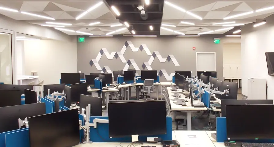 A modern office space for Raytheon with rows of desks and computer monitors arranged in an open layout, surrounded by bright overhead lights and geometric ceiling designs.