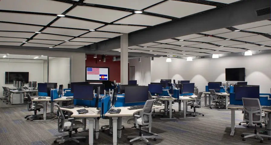 A large, modern office space of US Space Force TAPlab featuring rows of desks with computer monitors, ergonomic chairs, and blue dividers. The ceiling is fitted with rectangular acoustic panels and recessed lighting.